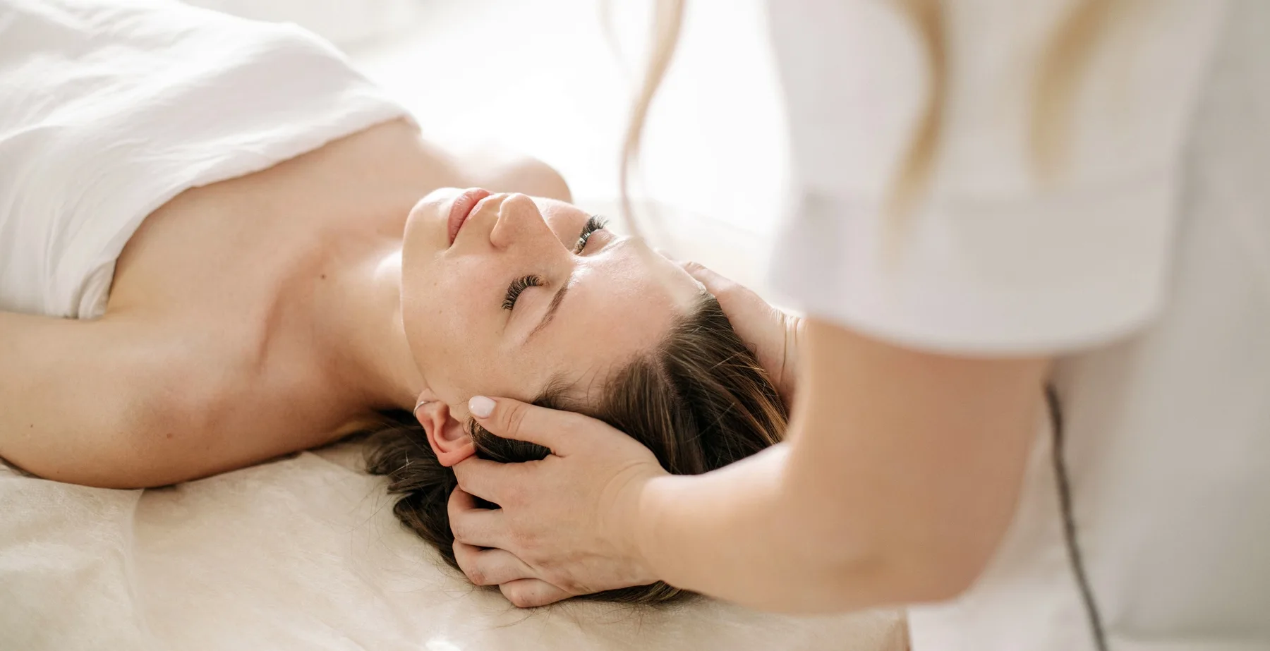 Žena leži zatvorenih očiju na masažnom stolu dok joj terapeut nežno masira glavu i lice tokom opuštajućeg facial tretmana.