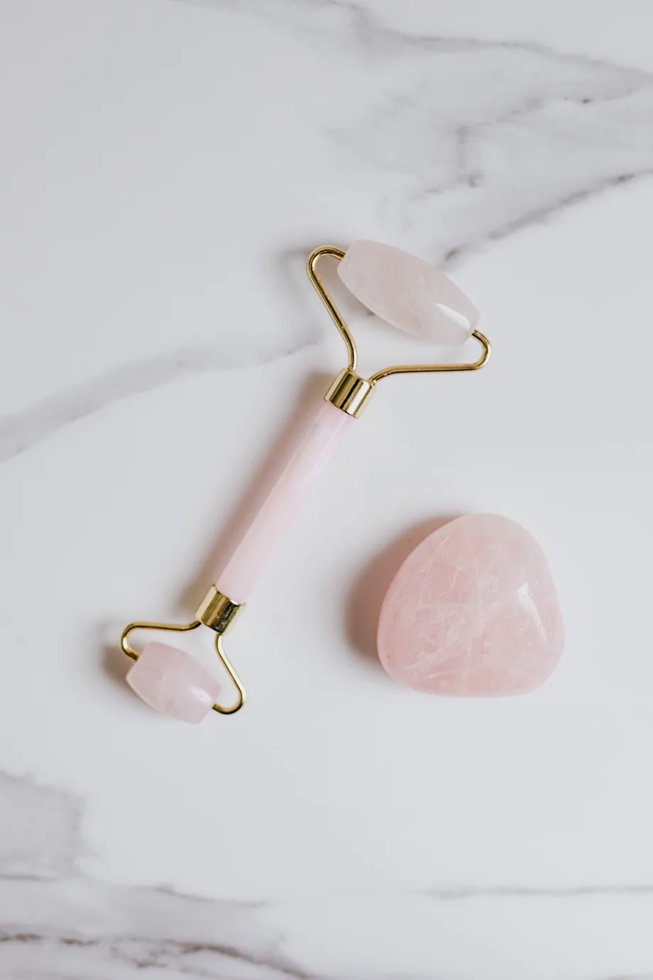 Roze quartz roler za lice i kamen za gua sha masažu položeni na svetloj mermernoj podlozi.