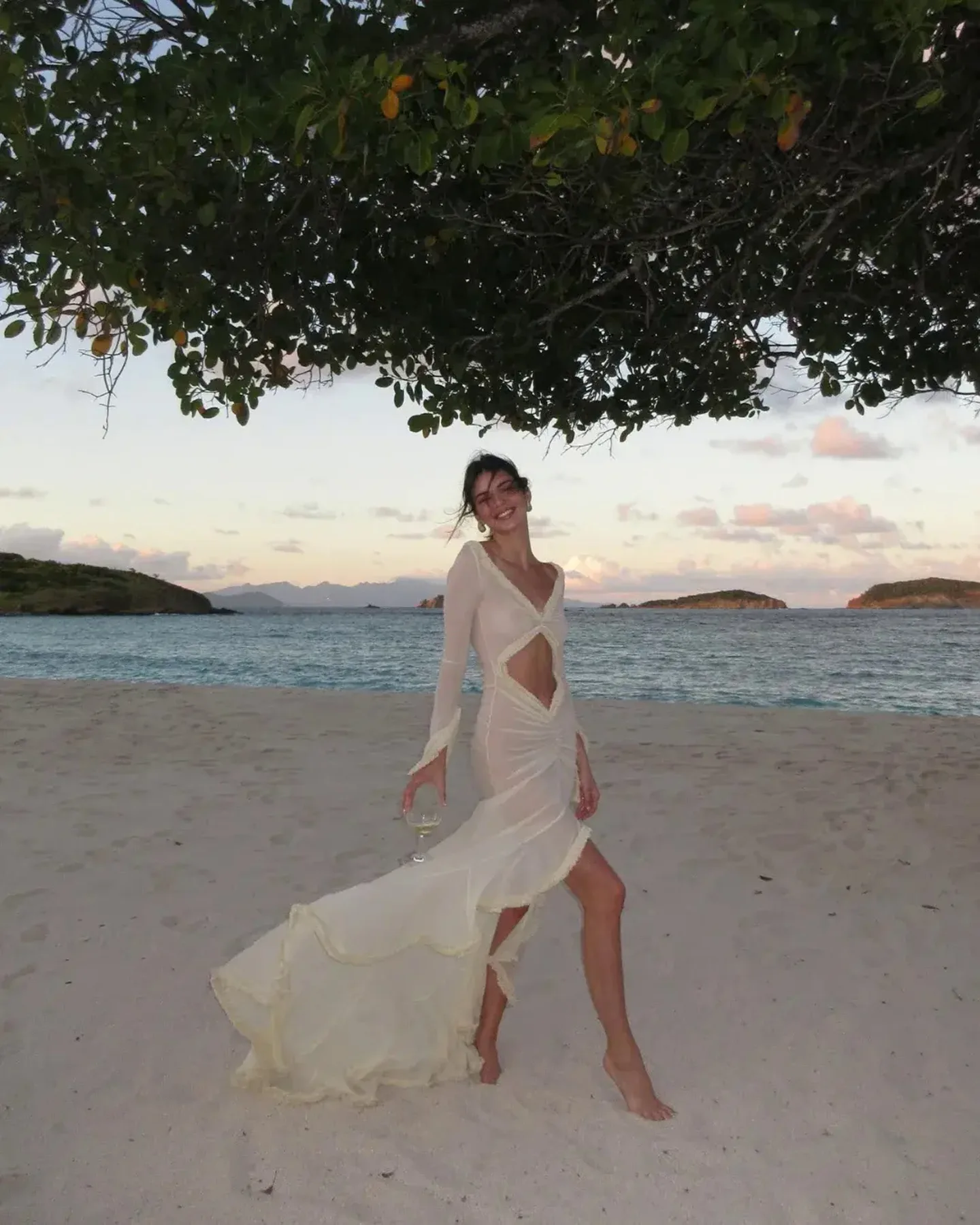 Kendall Jenner dugoj beloj haljini hoda bosonoga po peščanoj plaži, ispod krošnje drveta, uz more u pozadini.