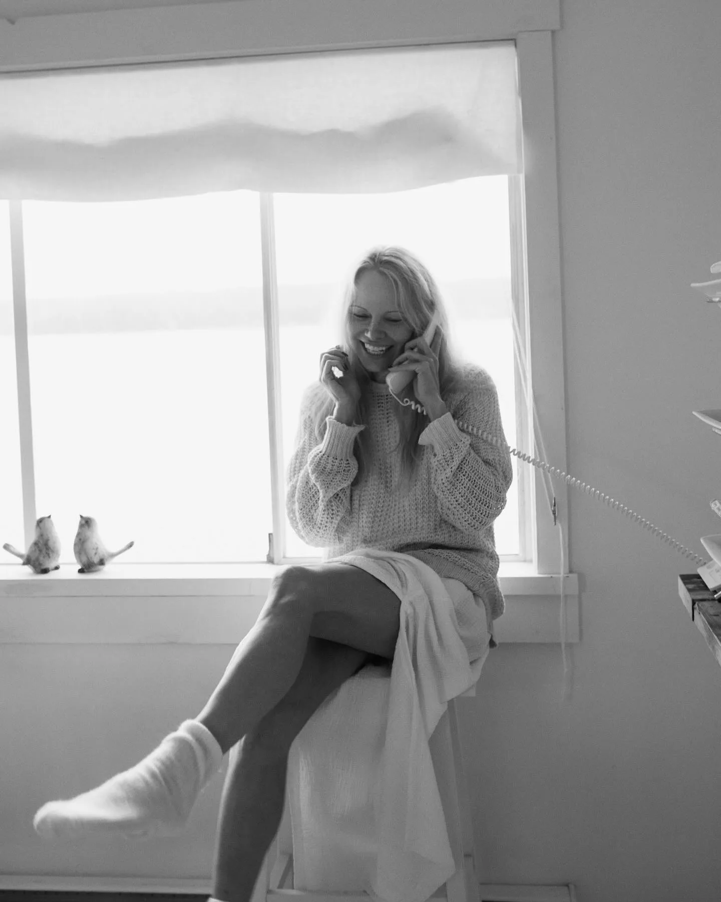 Nasmejana žena sedi pored prozora i razgovara telefonom.
