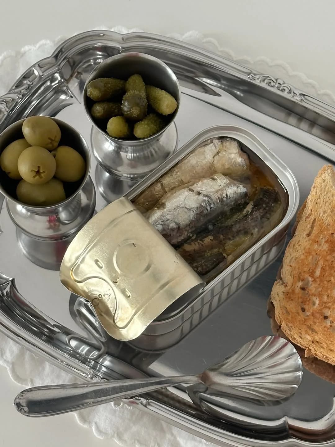 Otvorena konzerva sardina na srebrnom poslužavniku, uz zelene masline, kisele krastavčiće i tostirani hleb.