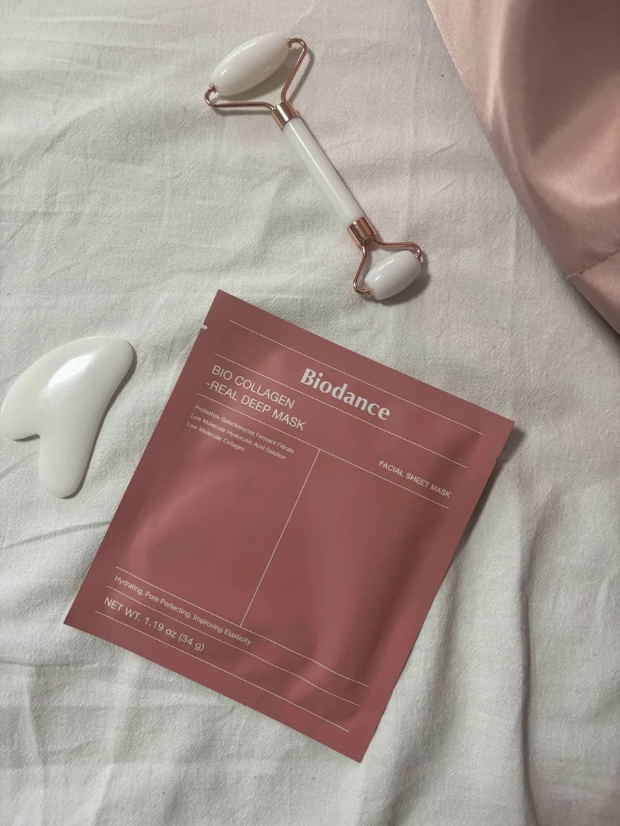 Biodance Bio Collagen maska za lice, roler i gua sha alat za masažu položeni na svetlu tkaninu uz detalj roze satena.